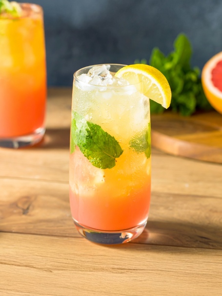 Un cocktail Green Label de couleur dorée avec un léger dégradé rosé, servi dans un verre long drink transparent rempli de glaçons. Le cocktail est décoré de feuilles de menthe fraîches et d’une fine tranche de citron posée sur le rebord du verre, offrant une présentation élégante et rafraîchissante.