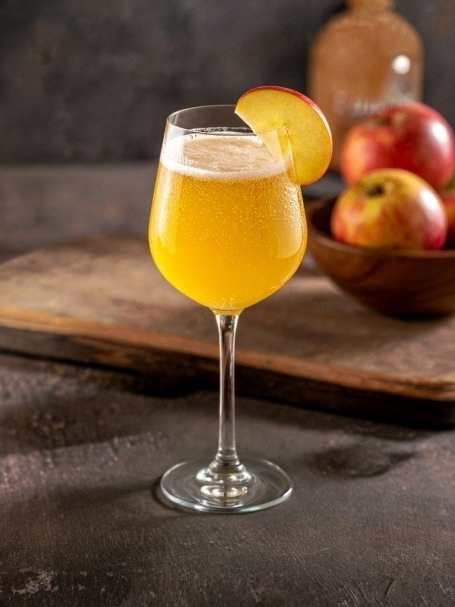 Cocktail Kir Barton, dans un verre à vin, à base de Whisky canadien Sam Barton, de jus de pomme, de prosecco Perlino Biologico avec une tranche de pomme.