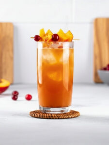 Cocktail Péché Mignon, dans un verre long drink, à base de Vodka Poliakov, de nectar de pêche, de jus de cranberry, de crème de pêche L'Héritier-Guyot et de glaçons, avec une brochette de fruits.