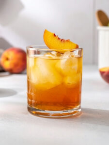 Cocktail Ti'Peach dans un verre old fahsioned, à base de Rhum VO Dillon, de Crème de pêche L'Héritier-Guyot, de Jus de citron vert pressé et de 2 tranches Gingembre, avec des glaçons et un morceau de pêche sur le rebord du verre.