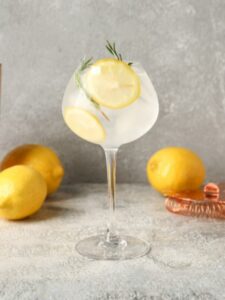 Cocktail Gin Tonic, servi dans un verre à pied, préparé avec du gin, du tonic et garni de rondelles de citron jaune et de branches de romarin.