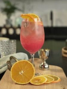 Cocktail Spritz sans alcool aux teintes orange vif, servi dans un verre piscine, décoré de rondelles d'orange dans un décor uni blanc avec des quartiers d'orange posées.