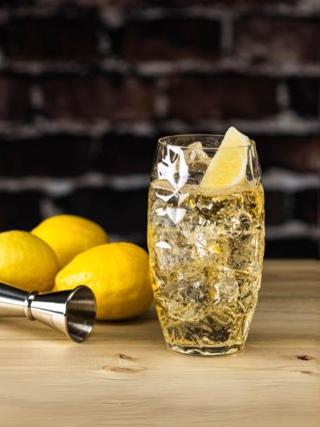 Cocktail whisky highball, préparé avec du blended scotch whisky et de l'eau gazeuse, servi dans un verre highball et garni d'une tranche de citron posé sur une table en bois avec des citrons jaunes et un doseur à cocktail