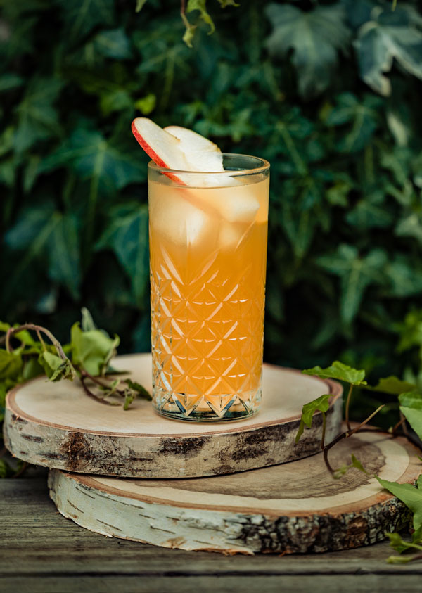 Cocktail Canadian Apple dans un long drink avec une tranche de pomme et de cannelle rondin de bois feuilles