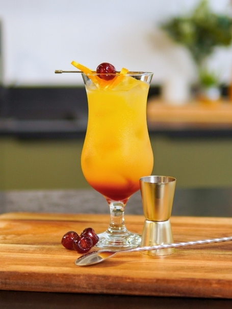 Cocktail sans alcool Afterglow, à base de jus d'orange, de jus d'ananas et de sirop de grenadine, dans un verre long drink, avec des glaçons.