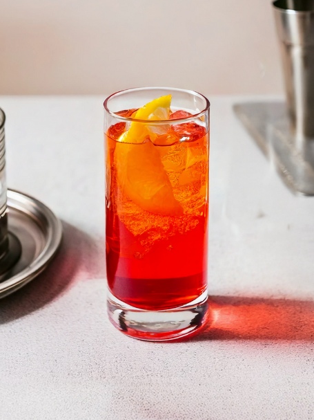 cocktail tampico de couleur rouge et orange vif servi avec des glaçons et un quartier de citron jaune dans un verre long drink