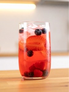 Verre tumbler posé dans une cuisine lumineuse et sur un comptoir en bois, rempli du mocktail fruits rouge et eau de coco, servi avec des fruits rouges (fraises, framboises et myrtilles) et des glaçons.