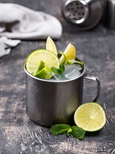 Cocktail London Mule servi dans un timbale, préparé avec du gin, du ginger beer et garni de rondelles de citron vert et de feuilles de menthe posé sur une table en bois gris