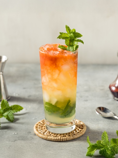 Cocktail Queens Park Swizzle servi dans un verre long drink avec de la menthe et des glaçons, laissant apparaitre un dégradé rouge et orange avec le bitter.