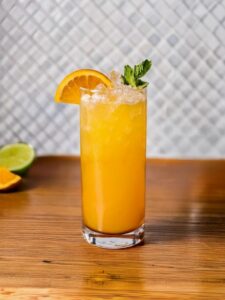 cocktail rum swizzle de couleur orange vif servi dans un verre long drink avec un quartier d'orange et une feuille de menthe en décoration sur le dessus du verre.
