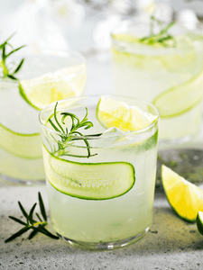 cocktail gin tonic avec des tranches de concombre et un quartier de citron jaune déposé dans un verre décoré avec une branche de romarin frais