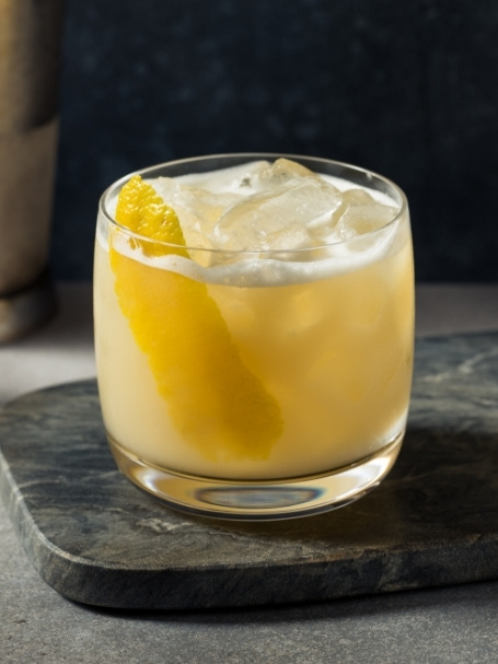 Cocktail Brandy Sour dans un verre old fashioned avec de la mousse, des glaçons et un zeste de citron jaune, à base de cognac, de jus de citron jaune, de sirop de canne et d'un blanc d'oeuf.