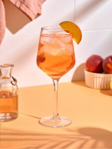 Cocktail Virgin Spritz Pêche à base de Pétillant sans alcool rosé, de sirop de pêche et d'eau gazeuse. Dans un verre piscine avec des glaçons.