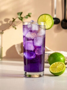 Cocktail Purple Rain dans un verre long drink à base de Vodka, de Curaçao bleu, de sirop de grenadine, de us de citron vert et de limonade, avec des glaçons et une tranche de citron vert sur le rebord du verre.