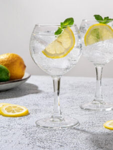 Verre ballon de gin tonic sans alcool rempli de glaçons et d’eau pétillante, garni d’une rondelle de citron et de feuilles de menthe fraîche. Un second verre similaire apparaît à l’arrière-plan, avec des citrons et un citron vert posés sur une table claire dans une ambiance lumineuse et rafraîchissante.