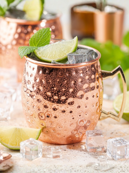 Deux mugs en cuivre contenant un cocktail sans alcool avec glaçons, quartiers de citron vert et feuilles de menthe, posés sur une table claire entourée de glaçons et de citrons verts frais.