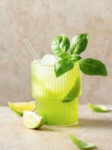 Verre transparent contenant un cocktail sans alcool vert clair avec glaçons, garni de feuilles de basilic fraîches. Le verre est posé sur une surface beige entourée de quartiers et moitiés de citron vert, de feuilles de basilic et d’une petite cuillère dorée. Fond neutre aux tons crème avec éclairage doux, mettant en valeur l’aspect frais et rafraîchissant de la boisson.