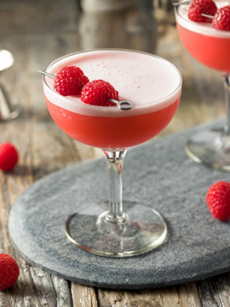 Cocktail Virgin Clover Club sans alcool à la framboise servi dans deux verres à cocktail, garni de framboises fraîches et d’une mousse légère, sur une table en bois rustique avec framboises fraîches et accessoires de bar.