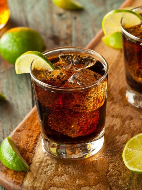 Deux verres de Virgin Cuba Libre remplis de glaçons sont servis sur un plateau en bois rustique. Les cocktails à la couleur ambrée sont garnis de quartiers de citron vert. Plusieurs citrons verts entiers et coupés sont disposés autour des verres sur une table en bois, créant une ambiance chaleureuse et rafraîchissante.