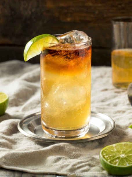 Virgin Dark 'n' Stormy, cocktail sans alcool rafraîchissant au gingembre et citron vert, servi sur glace dans un verre highball, avec citrons verts frais sur une table en bois au style authentique.