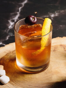 Cocktail Virgin Old Fashioned sans alcool servi sur glace dans un verre tumbler, garni d'un zeste d'orange et d'une cerise, présenté sur une tranche de bois avec jigger de bar et agrumes frais en arrière-plan.